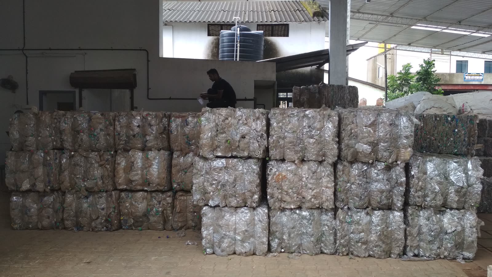 Stacked bales of clear PET bottles at a recycling facility with a worker seated behind them