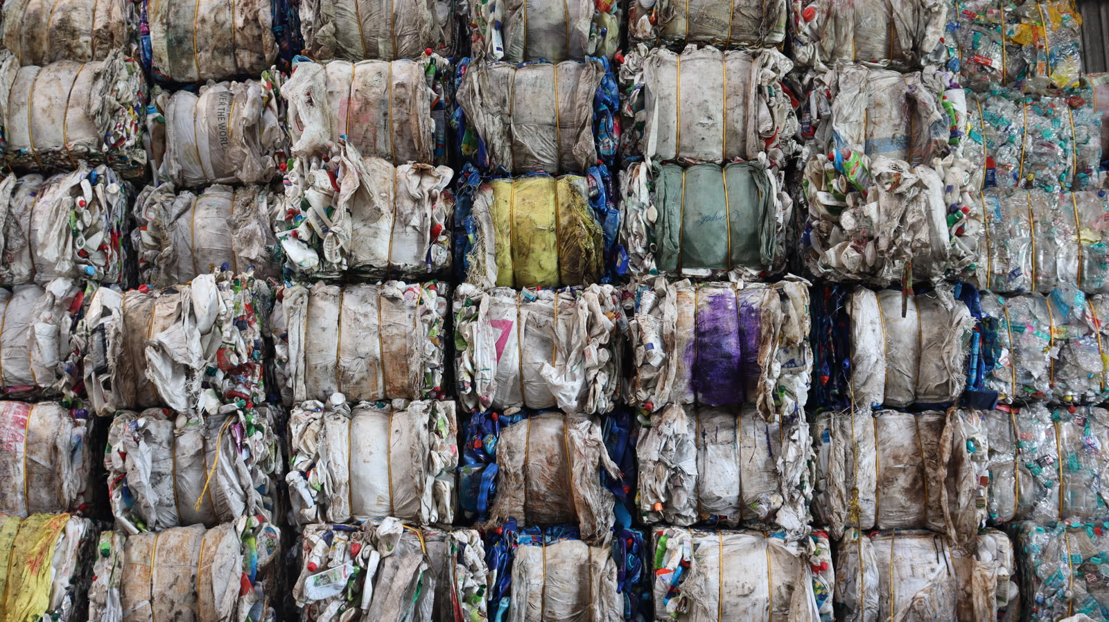 Stacked bales of mixed post-consumer plastic bound with yellow strapping, showing dirt, faded labels and bottle caps